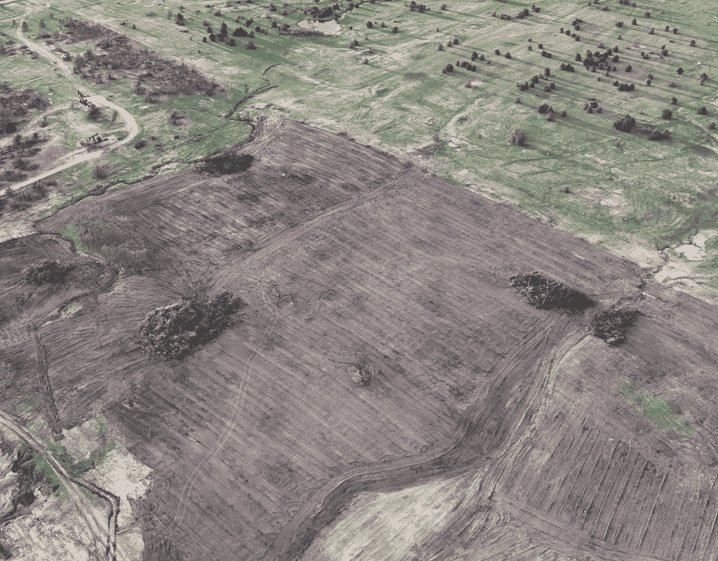 Aerial view of a large pasture in rural Oklahoma after machine clearing, showing rows of bare earth with scattered piles of brush and remaining trees on the perimeter.