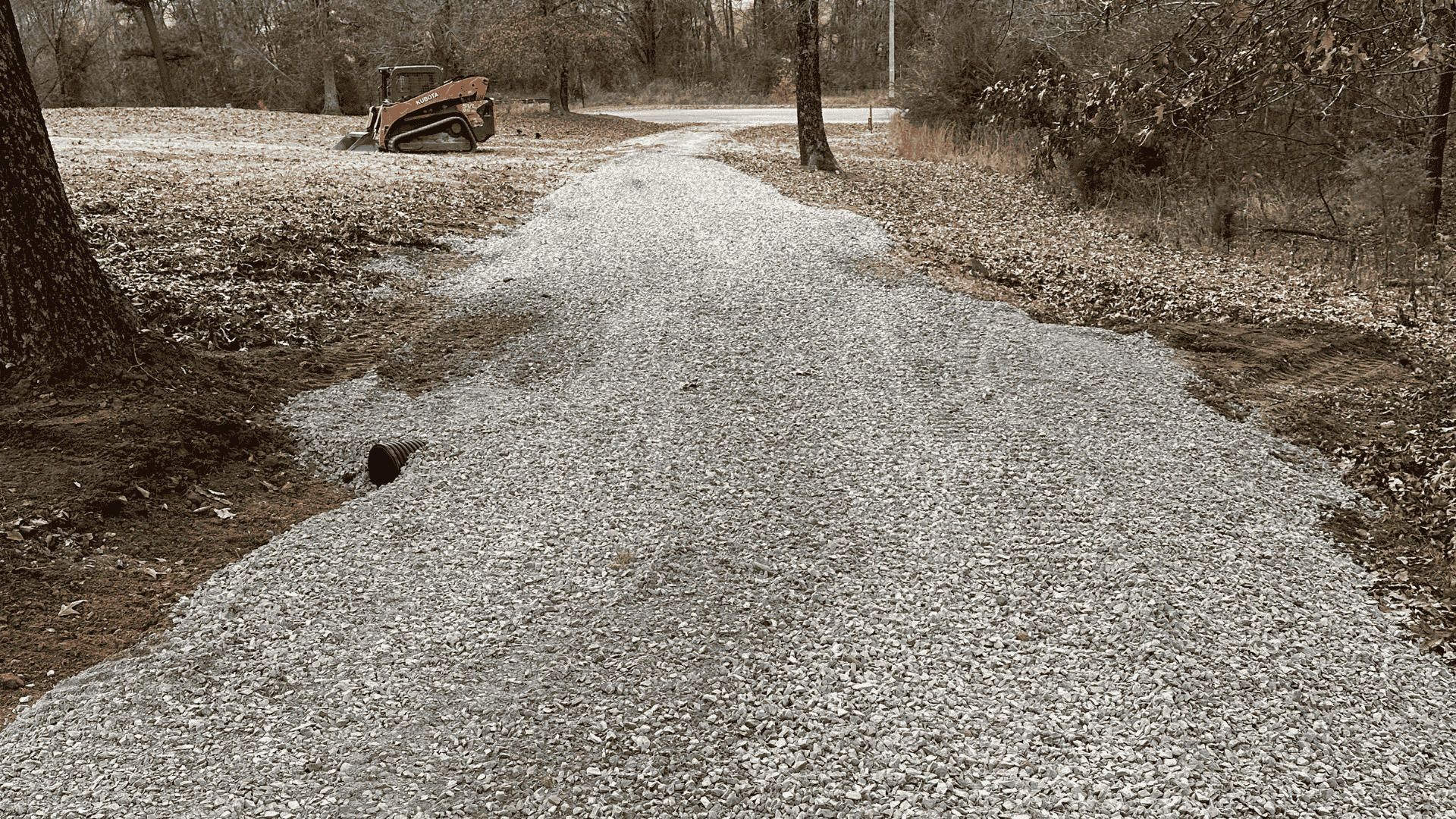 Gravel driveway freshly laid through wooded property, with a compact track loader in the background.