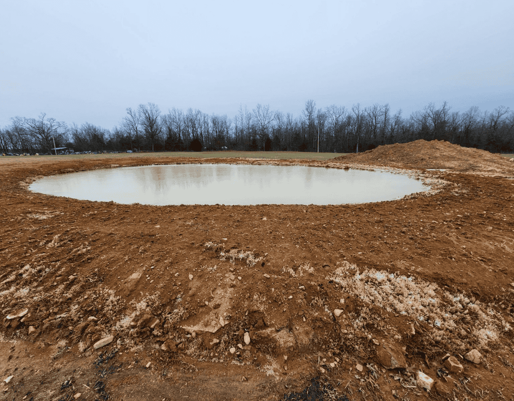 Close-up of a newly formed pond basin, showing smooth sloping banks of red clay and reflecting gray winter sky above.