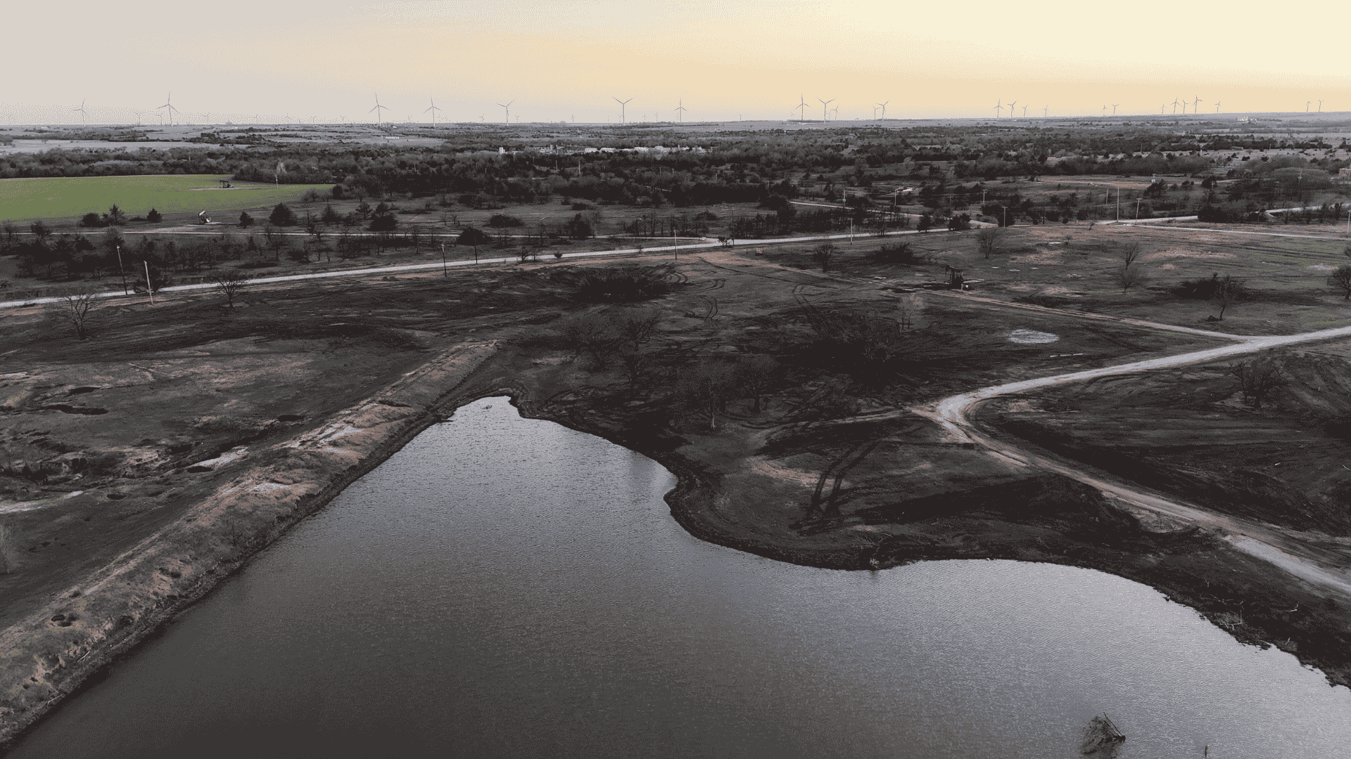 Overhead view of a newly dug pond basin in open pastureland, with smooth earthen banks and reflective water.