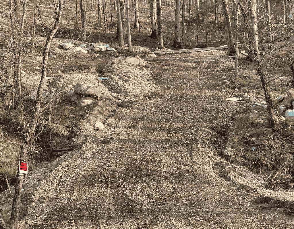 Bird’s-eye view of a leveled site pad amid a woodland edge, with gravel base and compacted red soil extending to standing trees.