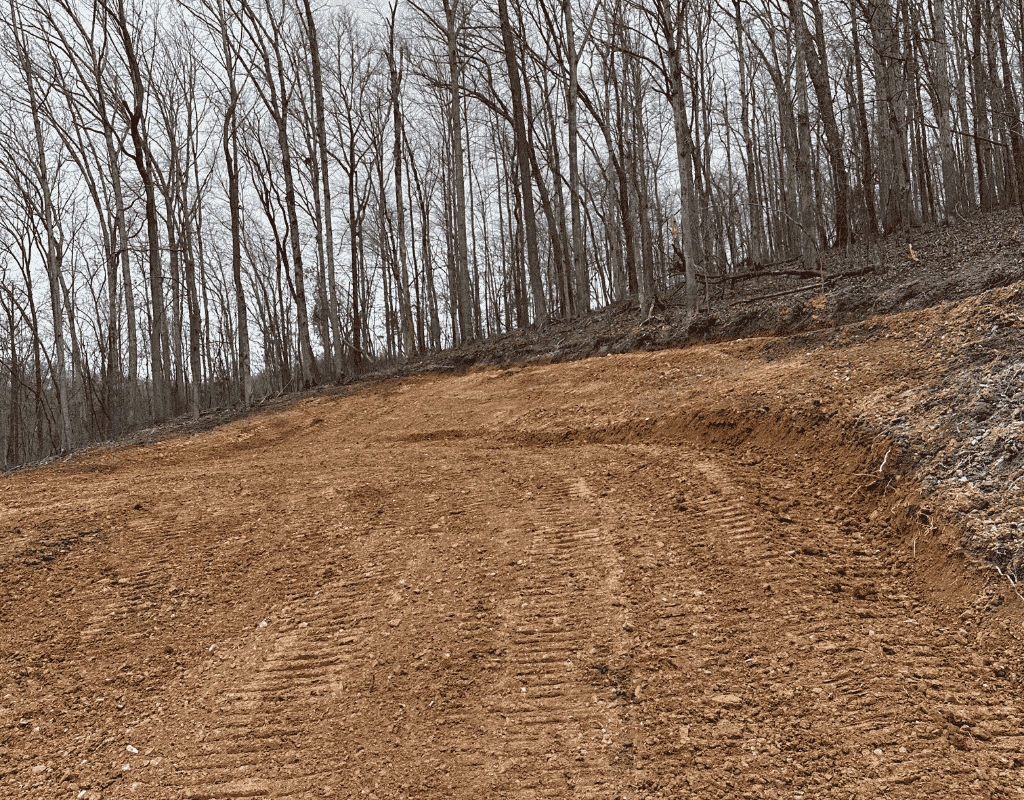 Ground-level image of a graded site pad surface, showing fresh earthwork edge and forest backdrop under an overcast sky.