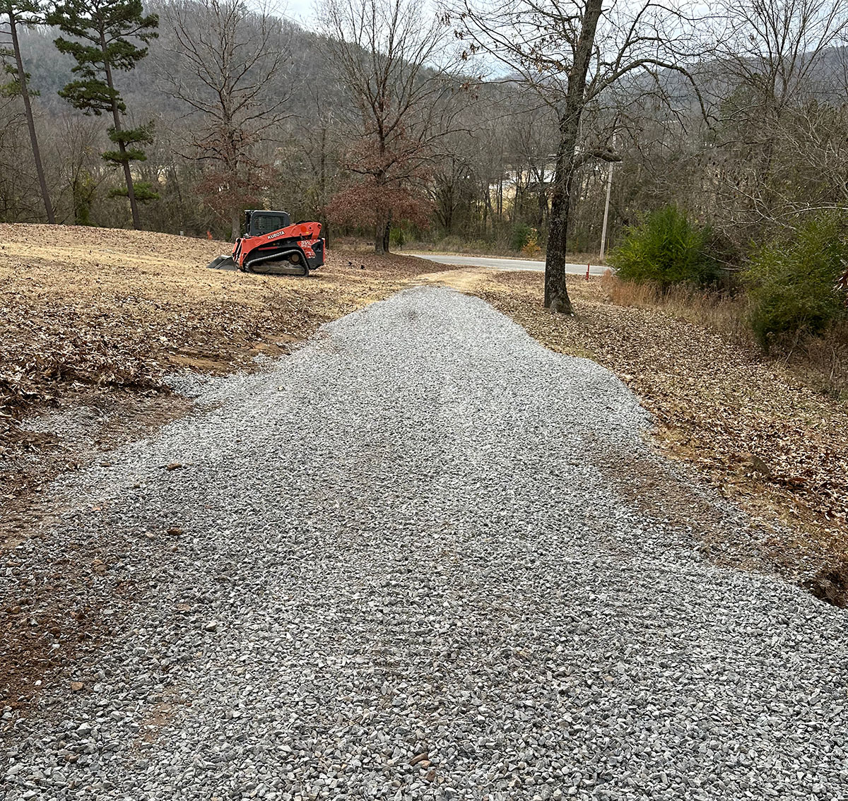 Fresh gravel over compacted soil at a rural home, with woods and a parked vehicle.