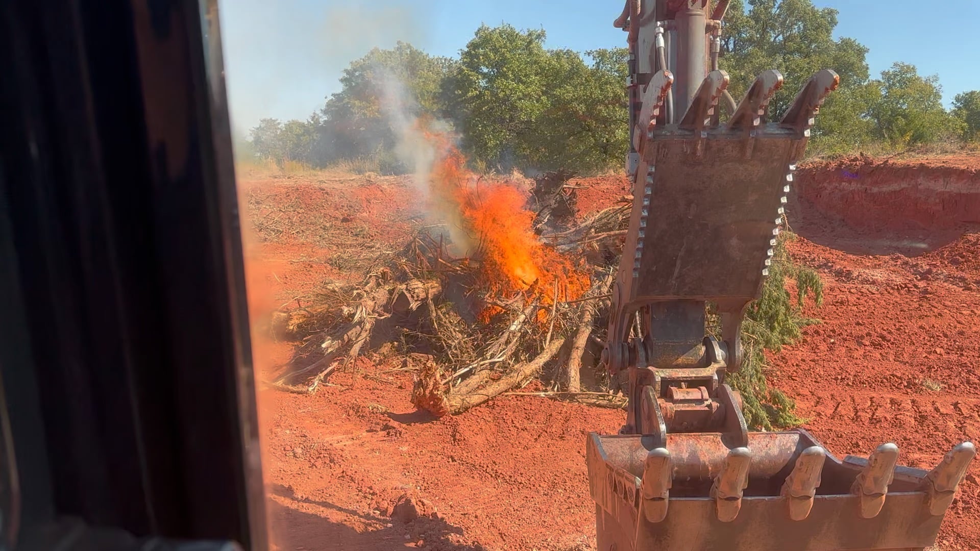 Excavator bucket near a brush fire on red dirt land.